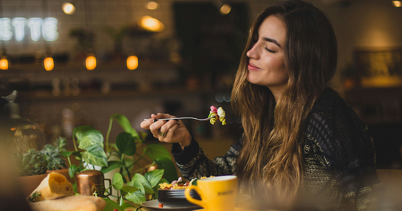 Woman mindfully enjoying a forkful of food at a table, pausing between bites as her natural fullness signals kick in